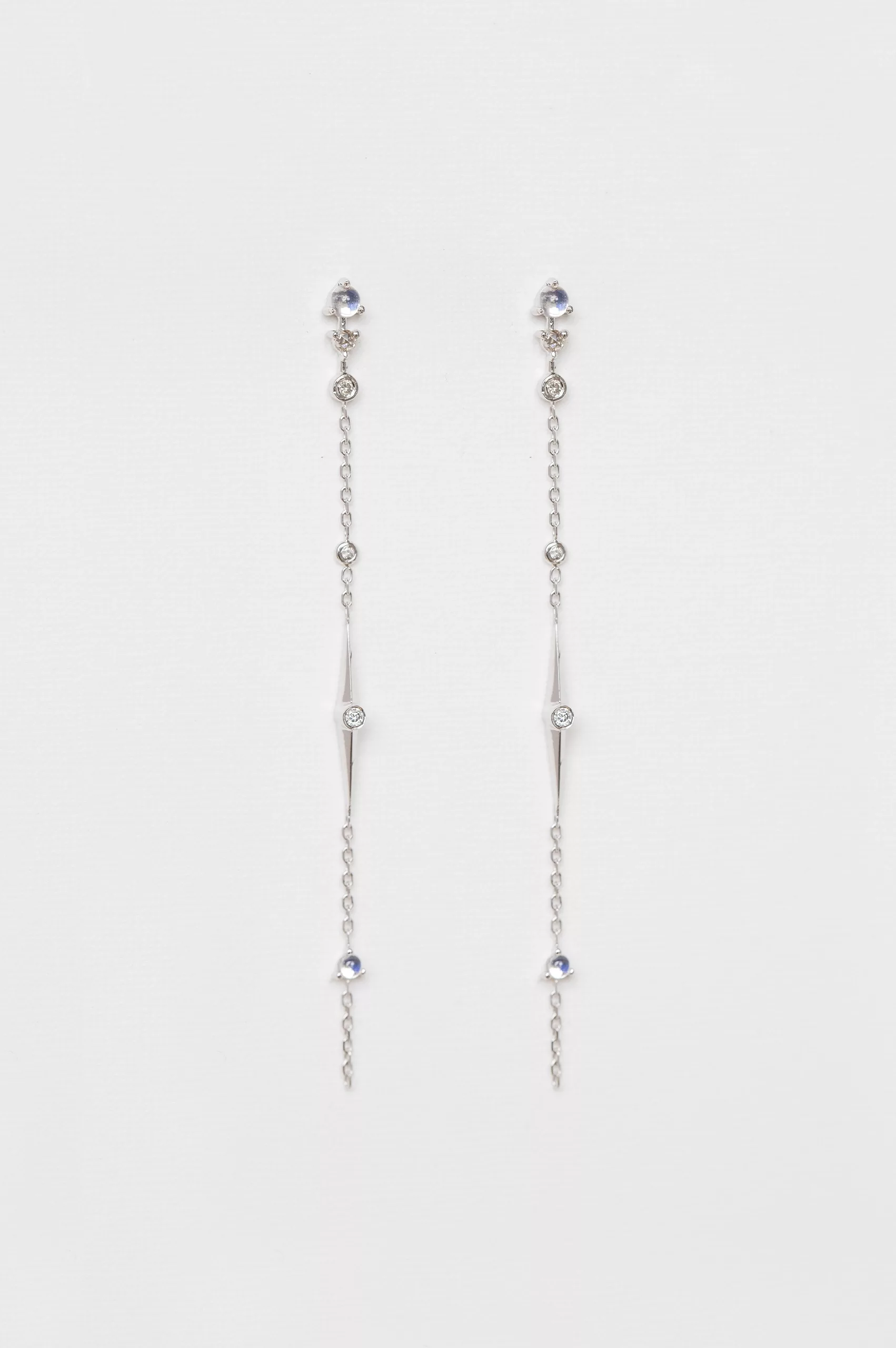 white gold long chain earrings each featuring a moonstone and diamond chain drop with dangling geometric details, worn as a delicate ear crawler